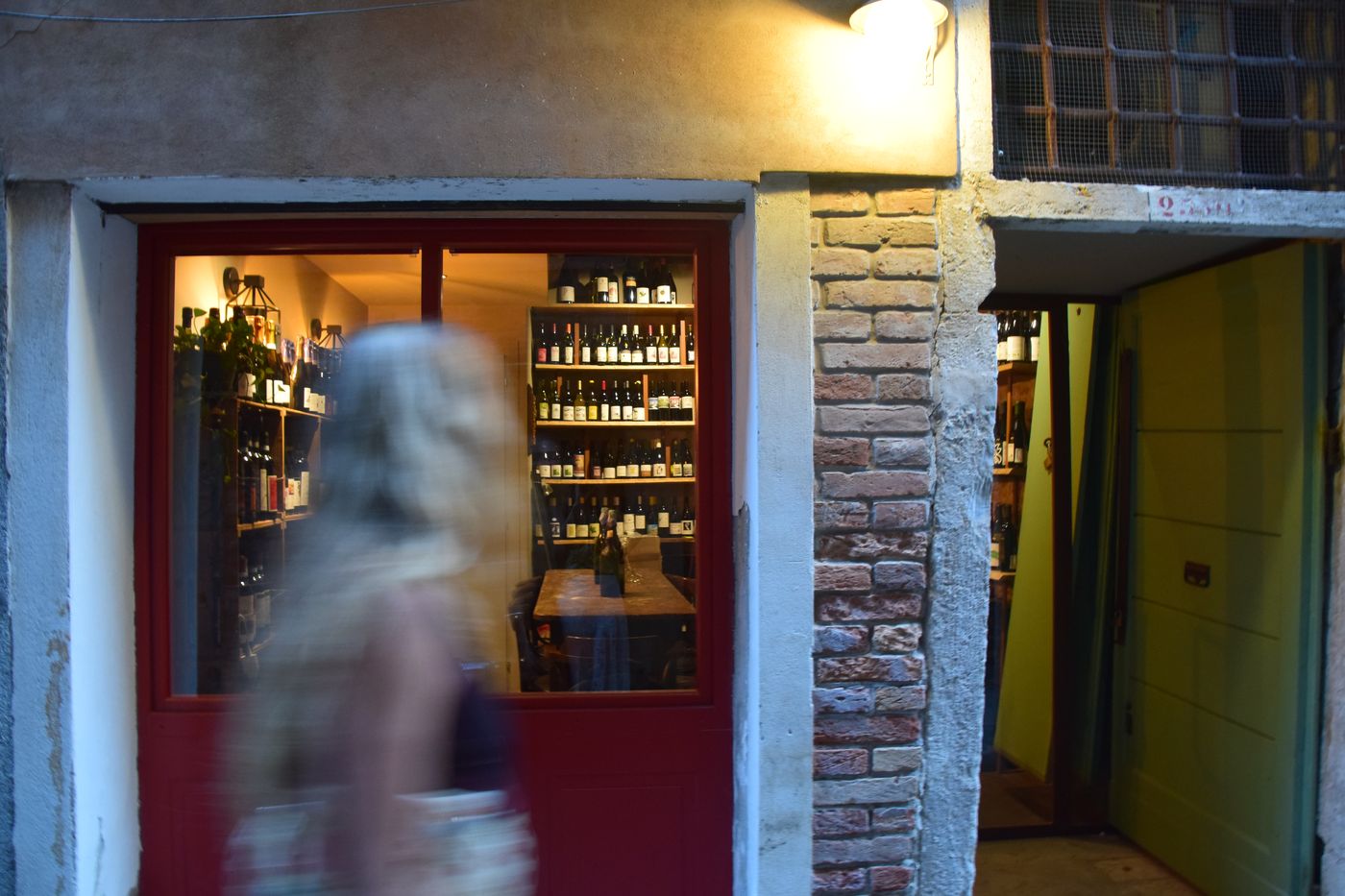 A lit wine bar seen from the street at night, red doorframe and shelves of bottles