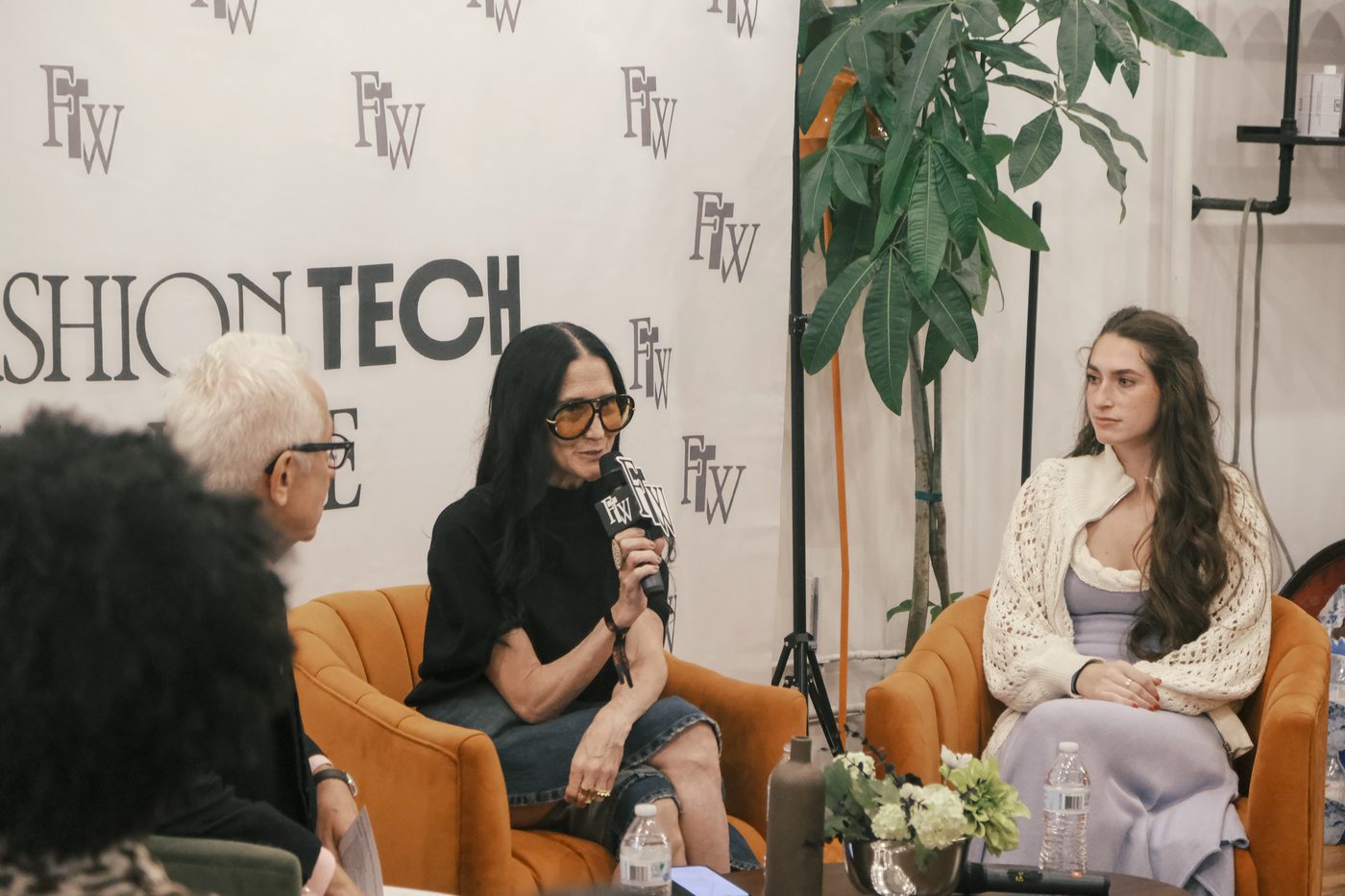 Three panellists seated in orange armchairs in conversation, one speaking into a branded microphone