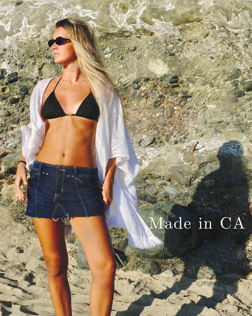 Campaign cover: a woman in a black bikini, open white shirt and dark denim mini standing against a rock face, the campaign line Made in CA set into her shadow