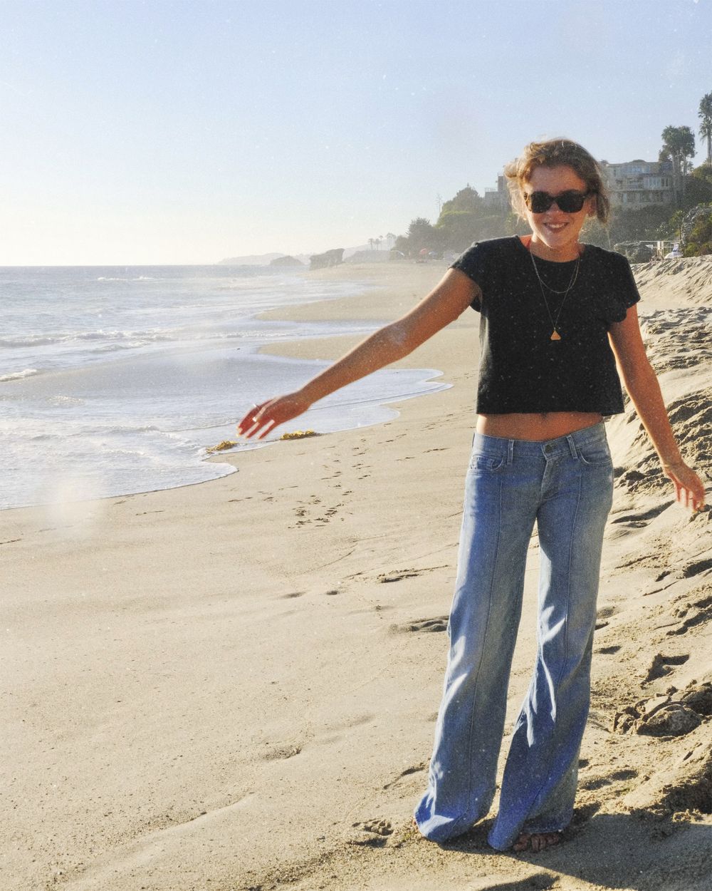 Standing on the shoreline in a black tee and light flares, one arm outstretched toward the water