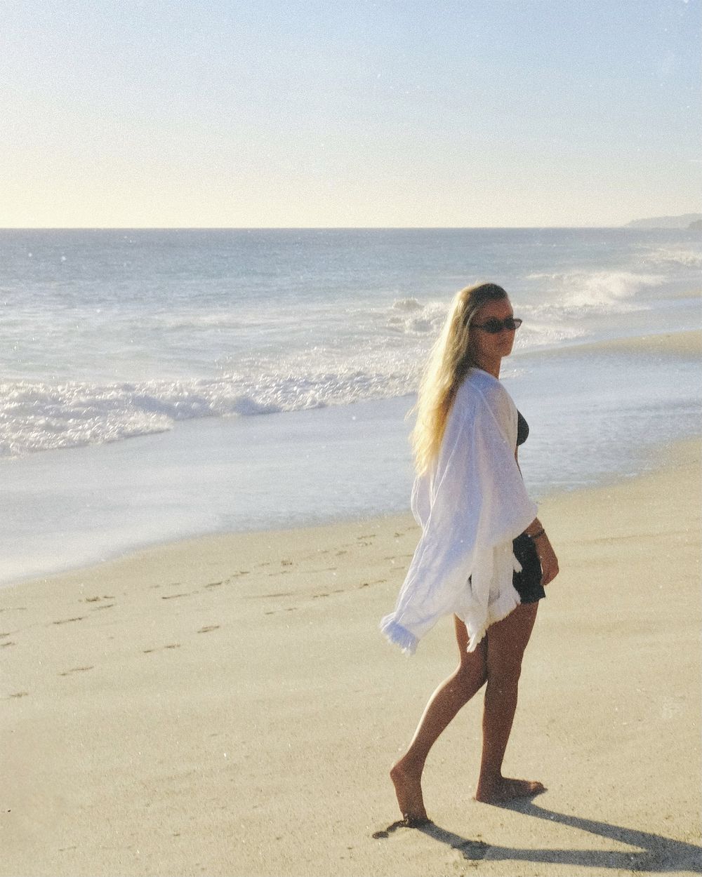 Walking away along the tideline in a white gauze cover-up over a black swimsuit