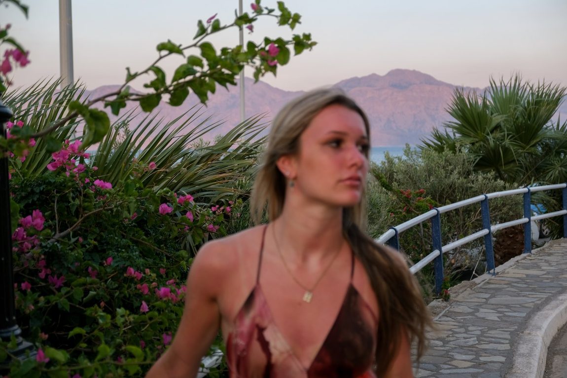 A woman looking out over a bay at dusk, bougainvillea and a mountain ridge behind her