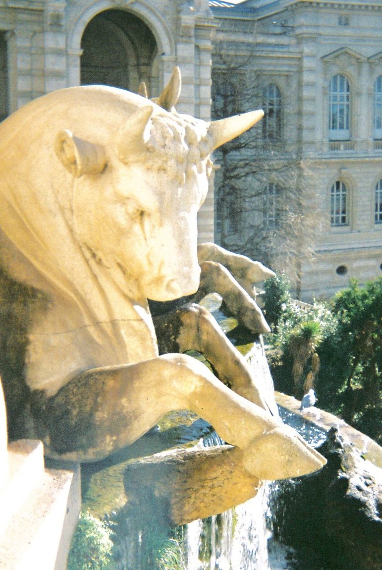 A gilded bull sculpture on a fountain, the stone facade of a palace behind it