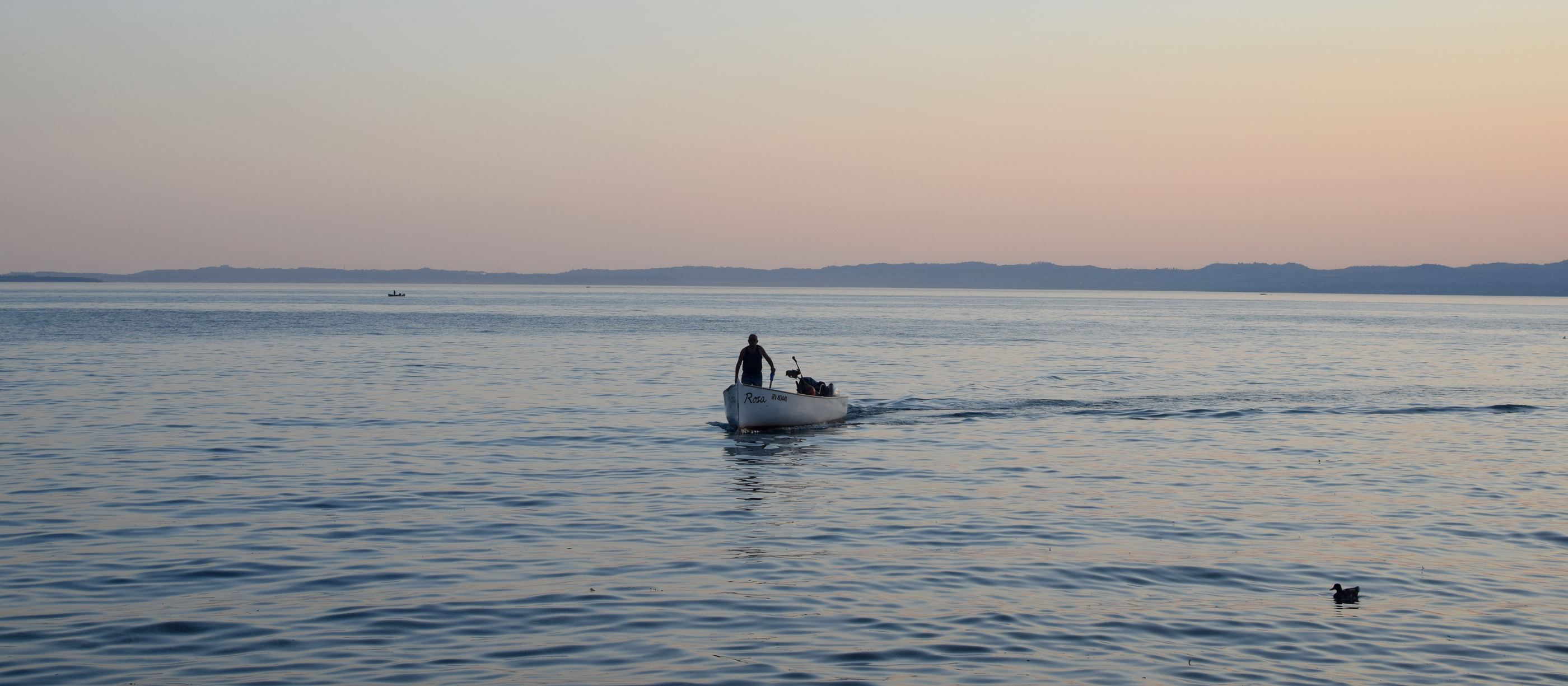 A fisherman steering a small boat across still water at sunrise, Lake Garda