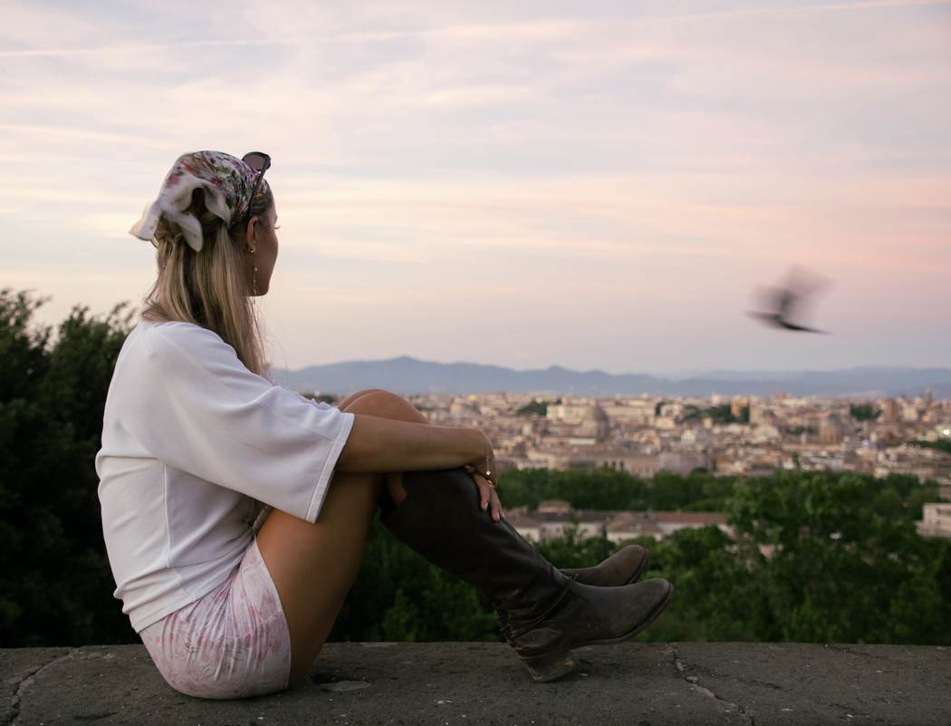 A figure seated on a wall above Rome at dusk as a bird passes
