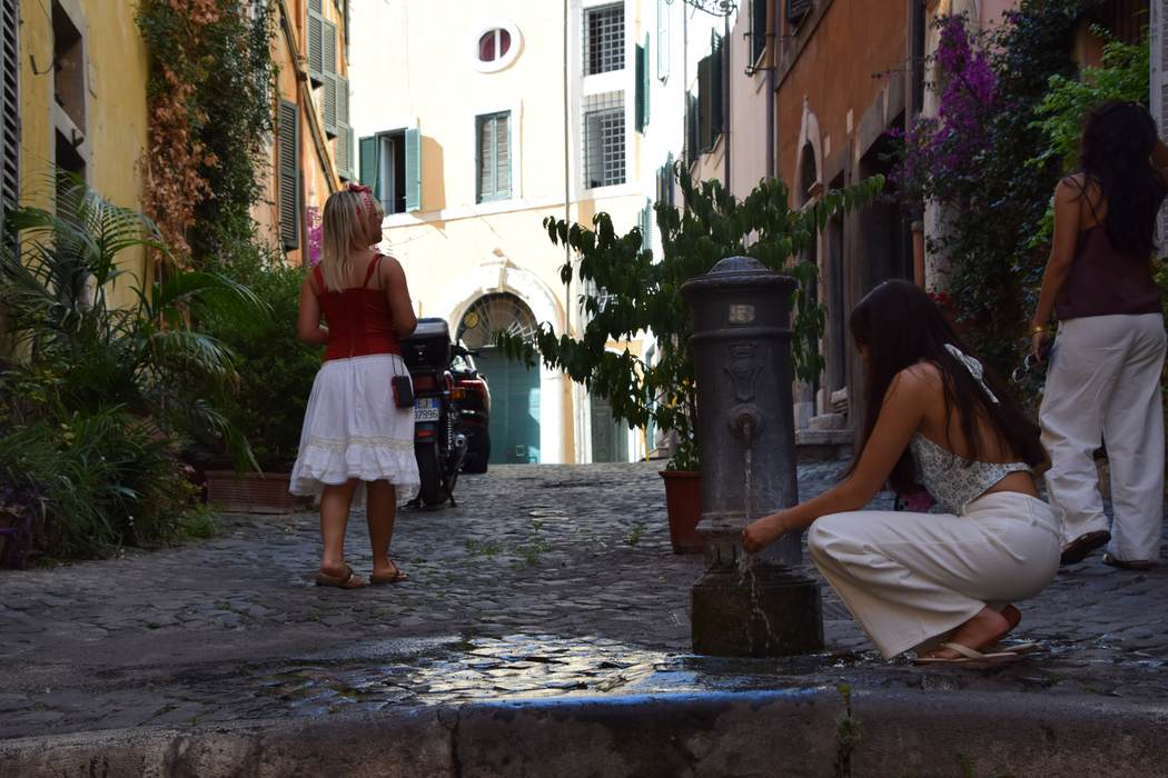 A cobbled Roman alley where someone crouches to drink from a street fountain