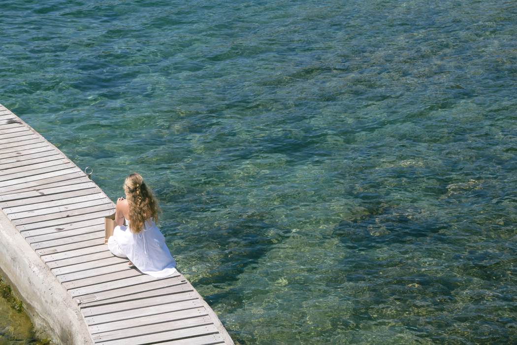 A woman in a white dress sitting at the end of a jetty over clear green water