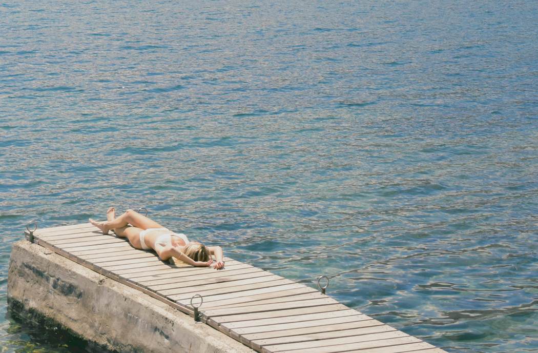 A figure sunbathing alone on a wooden jetty above blue water