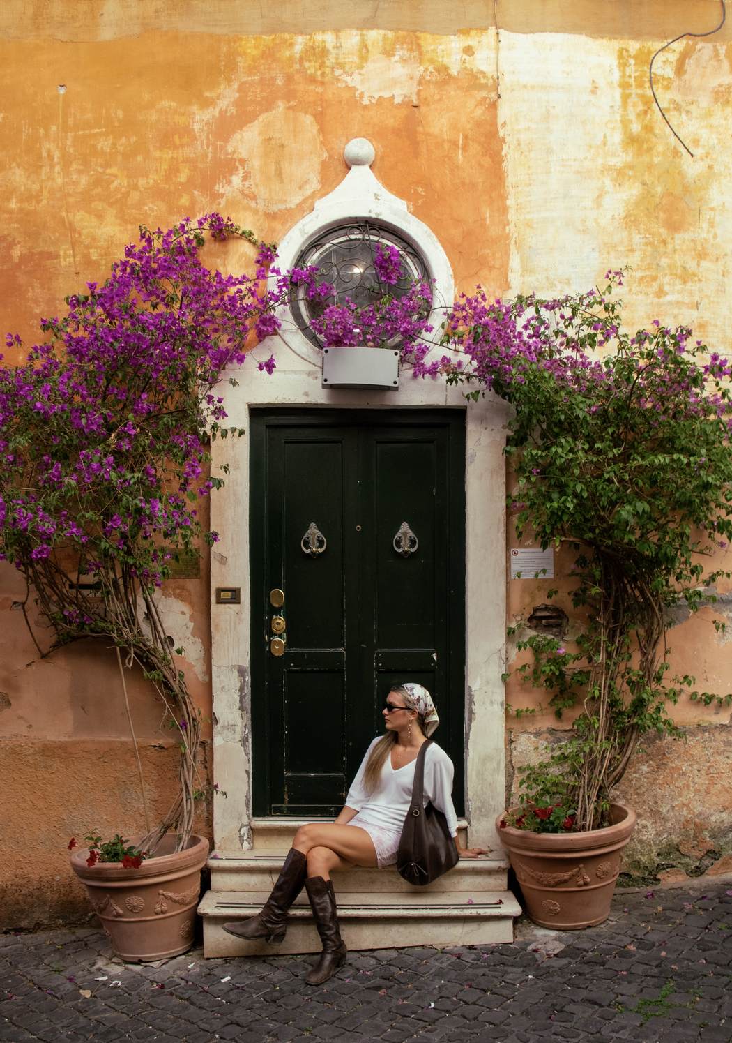 A figure seated on the step of a green door beneath cascading bougainvillea